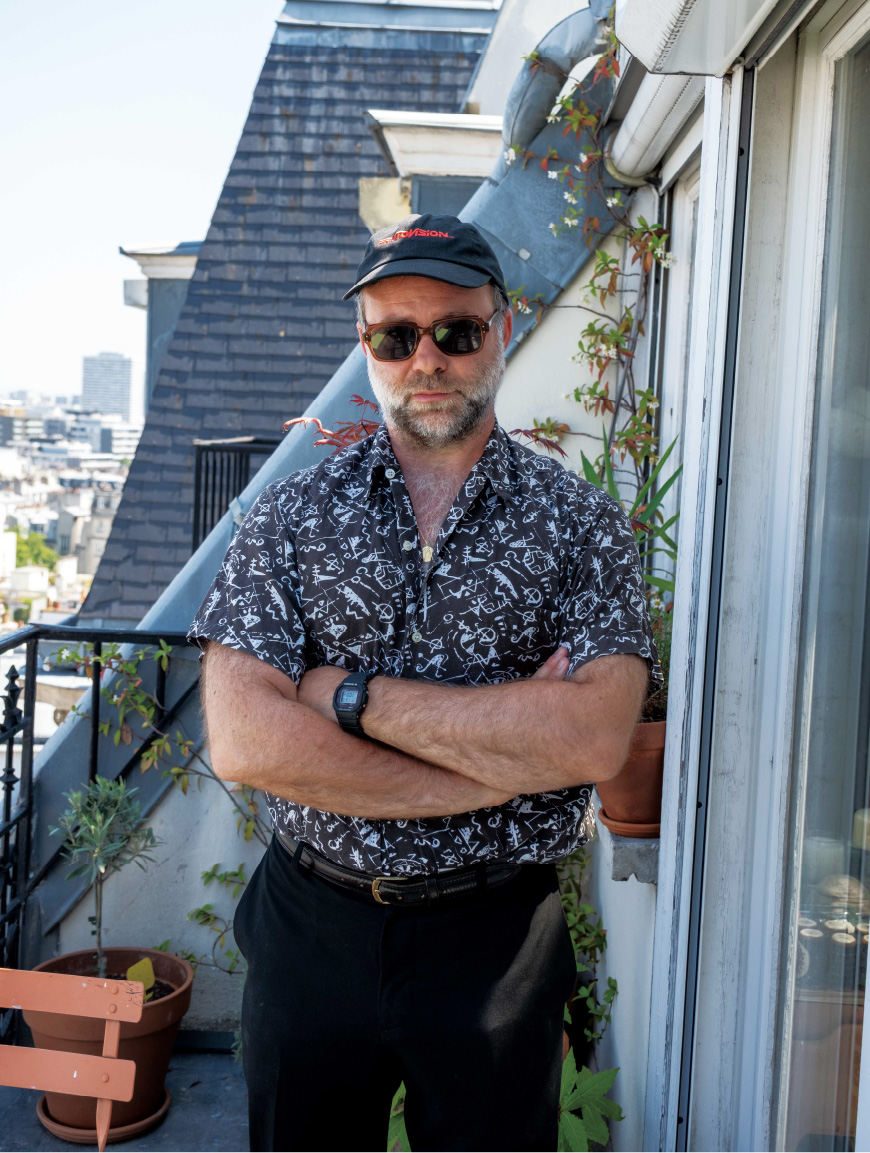 艺术总监头戴黑色帽子，身穿花纹衬衫，站在户外。